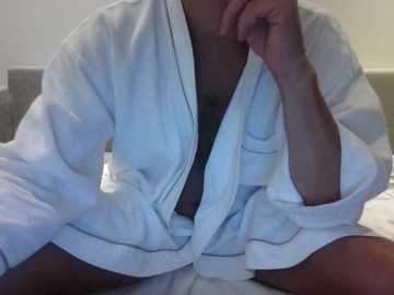 Video of a person in a white bathrobe, partially unzipped, revealing a black bra underneath. The individual is seated on a bed with a gray headboard in the background.