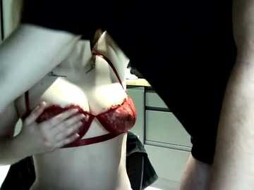 Video of a fair-skinned woman with large breasts in a red lace bra, being fondled by a person in black clothing. Background features a cluttered room with a white desk and scattered objects.