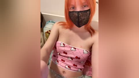 Video of a young woman with pale skin and orange hair, wearing a black face mask, pink Hello Kitty bandeau top, and white lace panties, sitting on a bed with a white headboard.