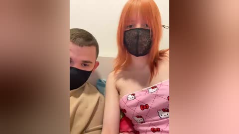 Video of a pale-skinned, orange-haired woman with a black face mask, wearing a pink Hello Kitty-patterned dress. Beside her, a short-haired man in a beige shirt and mask is partially visible.