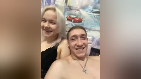 Video of a smiling blonde woman with fair skin and a man with a shaved head and fair skin, both shirtless, sitting on a bed. The background has a colorful, abstract mural.