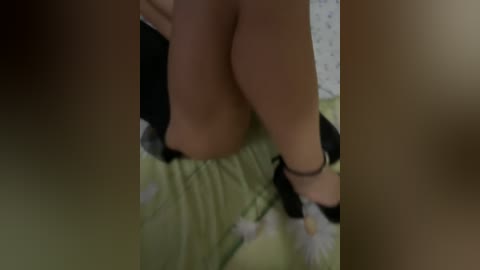 A blurred video showing a person with light skin, wearing black high heels and a floral-patterned green dress, seated on a bed with a polka-dotted pillow.