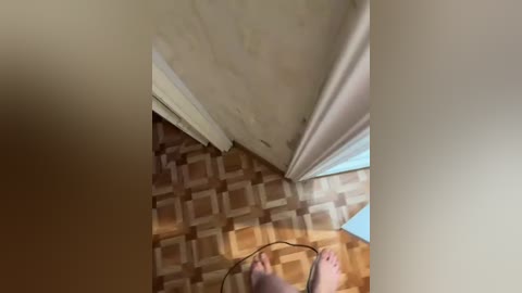 Video of a person with a white shirt, standing in a room with beige walls, a white ceiling, and a brown and beige patterned carpet. The person is holding a black cord, with their feet visible in the foreground.