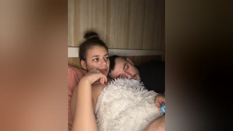 Video of a young woman with light skin and brown hair tied in a bun, resting her head on a man's chest. The man has dark hair and a beard, both in a cozy, dimly lit room.