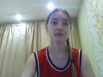 A video of a young woman with light skin and long blonde hair tied back, wearing a red basketball jersey. She stands in a room with beige curtains and a floral-patterned wall, under bright ceiling lights.