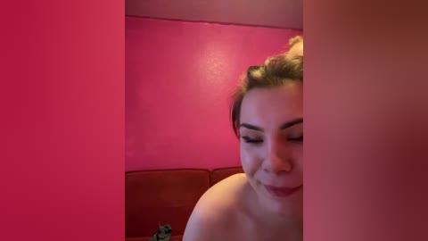 A video shows a smiling, light-skinned woman with dark hair tied back, standing in front of a pink wall, partially obscured by a vertical, translucent pink partition.