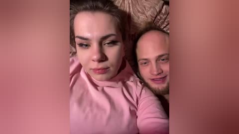 Video of a young woman with light skin and brown hair lying on a pink bedspread, looking serious, and a bald man with a beard smiling beside her.
