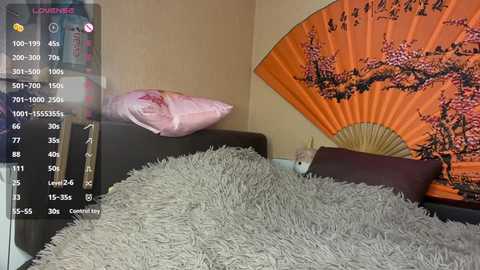A video of a cozy bedroom featuring a plush grey bedspread, a pink pillow, and a vibrant orange fan with cherry blossom design. A fluffy white cat rests on the bed, adding a soft texture.