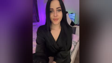 Video of a smiling, fair-skinned woman with long black hair, wearing a black satin robe, sitting on a bed with a TV and purple lighting in the background.