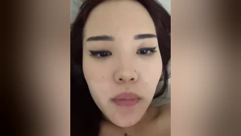 A close-up video of a young Asian woman with fair skin, wearing black eyeliner and minimal makeup, looking directly at the camera.
