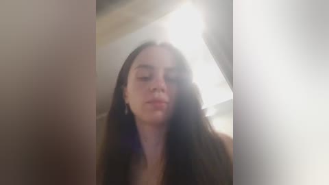 A blurry, low-resolution video of a young woman with long, dark hair, standing indoors. The image is slightly out of focus, with soft lighting and a bright spot on the right.