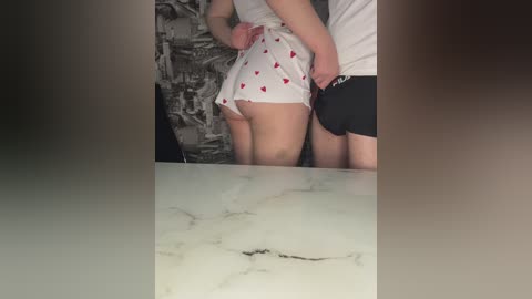 Video of a man and woman in a dimly lit room, both wearing white panties with red hearts. The woman has a light skin tone and a slim build.