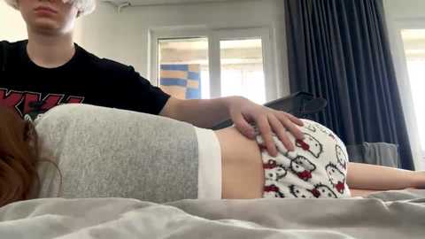 A video of a young woman with light skin and long brown hair, wearing gray pajama shorts with white waistband, lying on a bed. A young man with short white hair and a black t-shirt massages her back.