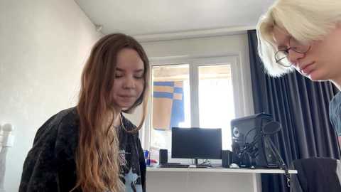 Video of a young woman with long, wavy hair and a woman with short, platinum blonde hair and glasses, both looking at a computer in a small, white-walled room with a window and a desk.