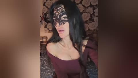Video of a woman with long black hair, wearing a black lace masquerade mask and a burgundy off-shoulder top, lying on a floral-patterned bed.