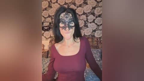 Video of a woman with fair skin, dark hair, wearing a maroon top, black lace mask, and floral wallpaper background.