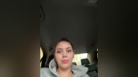 A video of a young woman with light skin and dark hair, wearing a grey hoodie, sitting in a car with dim lighting, looking slightly to the left.