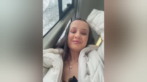 Video of a young woman with fair skin and straight brown hair, wearing a white puffer jacket, reclining in a car with a snow-covered landscape visible through the window.