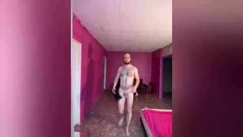 Video of a naked man with a muscular build, light skin, and a trimmed beard walking down a corridor with pink walls and a concrete floor.