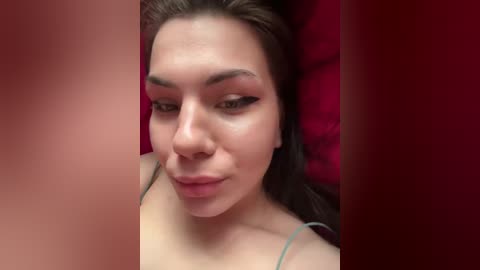 Video of a young woman with light skin, dark hair, and brown eyes, lying on a red pillow, wearing a green spaghetti strap top.