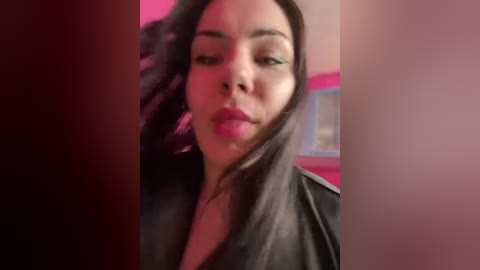 Video of a woman with medium skin tone, long dark hair, and full lips, wearing a black leather jacket. Background features a pink wall with a blurred painting.
