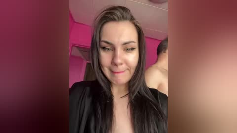 Video of a smiling woman with long black hair, fair skin, wearing a black robe, in a room with pink lighting and a ceiling fan.