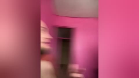 A blurry video of a person standing in a room with bright pink walls, wearing a white shirt and black pants. The image is heavily pixelated and out of focus.