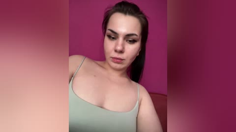 Video of a young, fair-skinned woman with dark hair, wearing a light green tank top, standing against a vibrant pink background. She has a neutral expression.