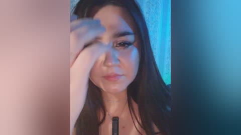 A close-up video of a young woman with long dark hair, light skin, and wearing a black top, shielding her face with her hand, against a blurred, light-colored background.