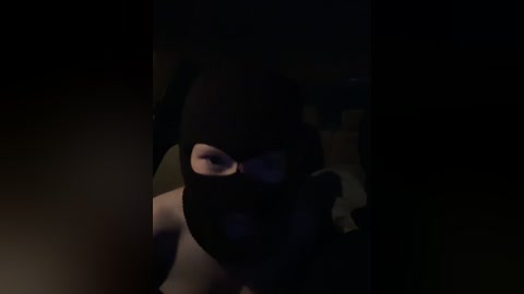 A video of a person wearing a dark, face-concealing mask, with their eyes visible, taken in a dimly lit room, emphasizing the shadowed and mysterious atmosphere.