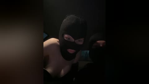 Video of a light-skinned woman in a black balaclava, wearing nothing but the mask, revealing her bare breasts, in a dimly lit, dark room.