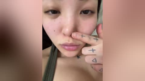 A close-up video of a young Asian woman with fair skin and dark hair, wearing a green top. She has a mole on her cheek and crosses her fingers, revealing tattooed crosses and triangles on her hands.