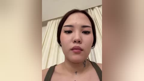 Video of a young Asian woman with light skin, wearing a green top, standing indoors. She has straight dark hair, black eyeliner, and a small cross tattoo on her neck. Cream-colored curtains are in the background.