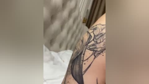 A video of a person's shoulder tattoo, featuring intricate black ink lines and dots, set against a blurred background of white sheets and a wooden headboard.