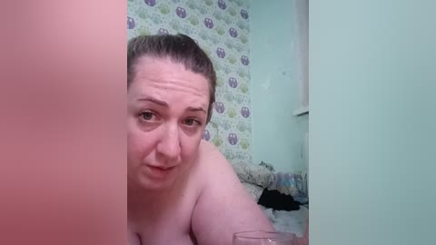 Video of a plus-sized, fair-skinned woman with brown hair tied back, looking over a partition, in a bathroom with patterned wallpaper and cluttered sink area.