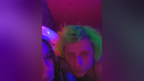 Video of a person with vivid, multicolored hair (green, yellow, and blue) and a serious expression, partially obscured by a glowing, multicolored light source.