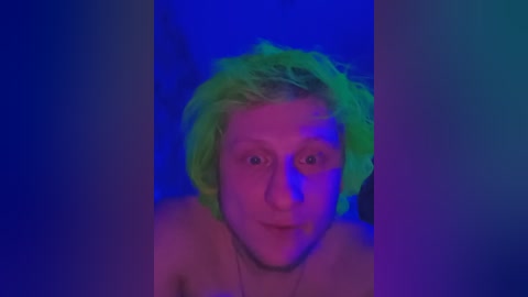 A video of a person with short, bright green hair, wide eyes, and a neutral expression, taken against a vivid blue background. The image is soft and slightly blurred, creating a surreal, dreamlike atmosphere.