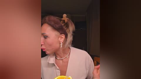A video shows a middle-aged woman with light skin and blonde hair in a bun, wearing a white blouse and pearl necklace, holding a yellow object, against a blurred, dark background.
