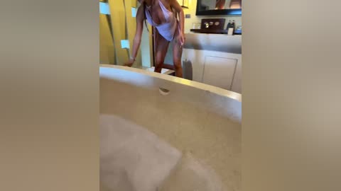 A video of a woman in a white tank top, bent over a bathtub, with a reflection in the mirror above. The background shows a modern bathroom with a glass shower enclosure and wooden cabinets.