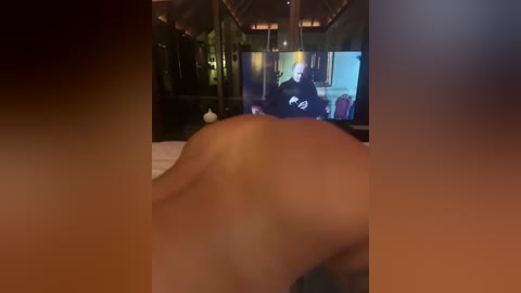 A video of a topless person lying on their stomach, with a blurry background showing a TV displaying a man's face. The room has wooden walls and a dim, warm light.