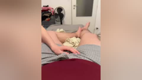 Video of a woman lying on a bed, wearing a gray shirt and maroon pants, with a man's hand on her stomach, in a cluttered, dimly-lit bedroom.
