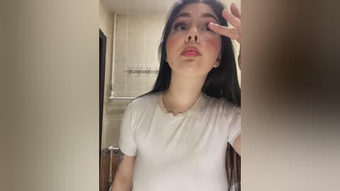 Video of a young Asian woman with long black hair, fair skin, and red lipstick, wearing a white t-shirt, in a beige-tiled bathroom, with a hand on her forehead.