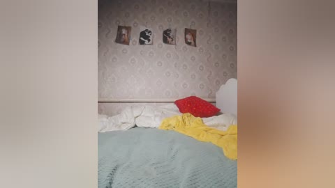 A video of a cluttered bedroom with a messy bed, featuring a yellow blanket, a red pillow, and a white sheet. The wallpaper is patterned, and three framed pictures hang on the wall.