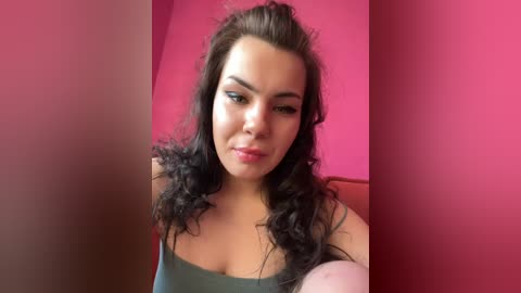 A video of a young woman with light skin and curly black hair, wearing a dark green sleeveless top, against a vibrant pink wall.