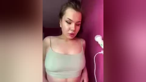 Video of a young Caucasian woman with light skin and brown hair tied back, wearing a light green sports bra, leaning against a pink wall, with a lamp emitting a soft glow.