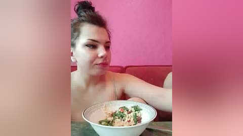 A video of a fair-skinned woman with dark hair in a messy bun, wearing a white top, sitting on a red couch against a pink wall. She holds a white bowl filled with a green salad and pasta.
