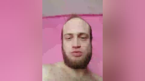 A video of a bald, bearded man with a neutral expression, wearing a pink shirt, against a pink background. The image is grainy and pixelated.