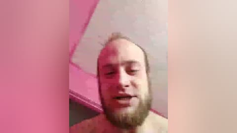 Video of a balding, bearded man with fair skin, looking introspective, against a pink-lit, textured ceiling.