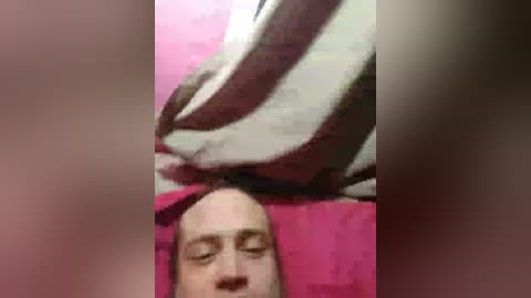 Video of a bald man lying on a pink towel, with a white and brown pillow behind his head, eyes closed, possibly asleep.