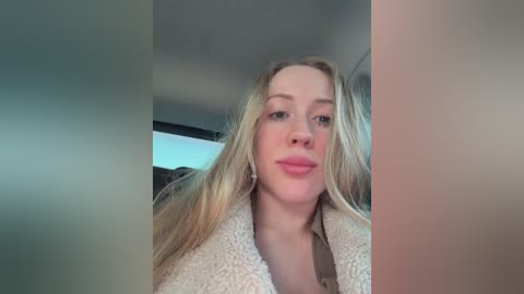 Video of a blonde woman with fair skin, wearing a beige cardigan, taken from a low angle, showing her upper body and face. Background is blurred, indoors.